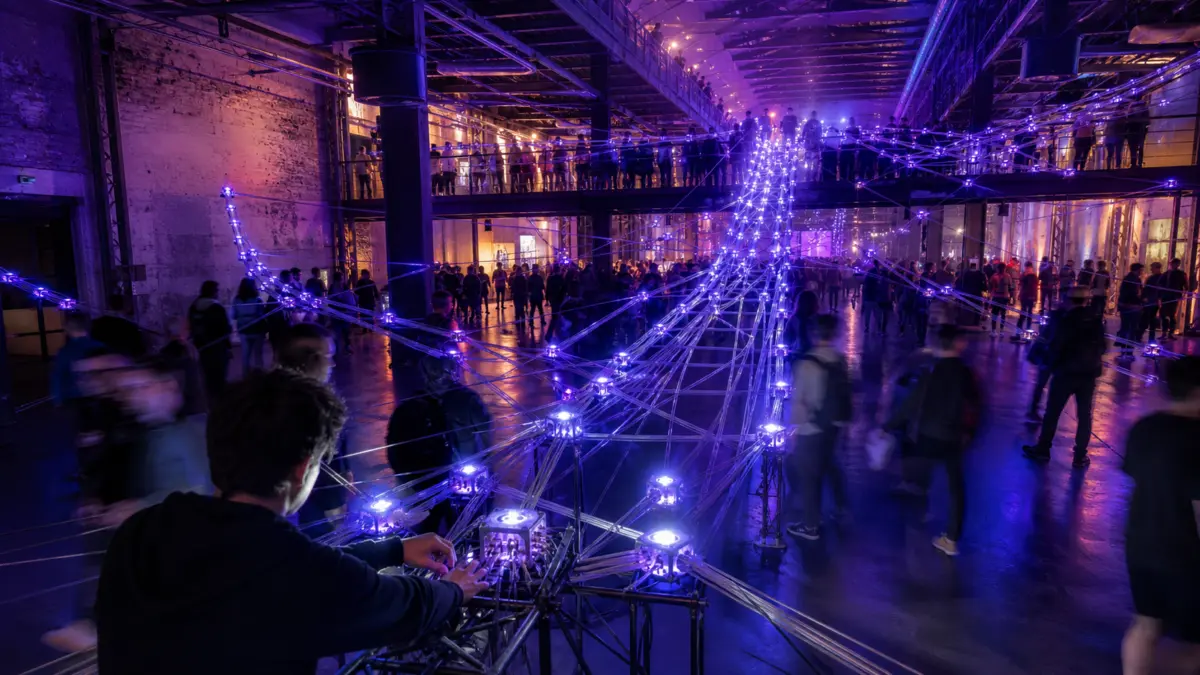 Anonyme Besucher bewegen sich durch eine verzweigte violett-blaue Netzwerkinstallation in einer Industriehalle